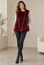 Load image into Gallery viewer, A woman models a sleeveless burgundy faux fur vest with a silver zipper over a gray long-sleeve top, black skinny jeans, and black ankle boots in a modern, neutral-toned living room setting.
