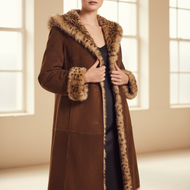 A woman modeling a long brown suede-like coat with a thick tan and brown fur-trimmed collar and cuffs, worn over a black dress in a bright, minimalist room with large windows.