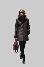 Įkelti nuotrauką į galerijos peržiūros programą, A woman modeling a dark brown leather coat with a contrasting black collar, styled with black pants, black boots, sunglasses, and a burgundy handbag, set against a solid gray background.
