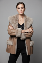 Load image into Gallery viewer, A model wearing a tan faux-leather jacket with a thick beige fur collar, cuffs, and pockets, styled over a black satin top and black trousers, against a solid gray studio background.
