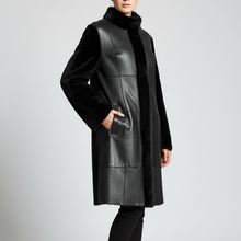 Įkelti nuotrauką į galerijos peržiūros programą, A model wearing a black faux-leather and fur coat with a high collar, black pants, and black high-heeled boots, standing against a light gray studio background.
