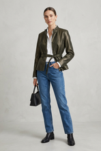 Load image into Gallery viewer, A woman stands against a light gray studio background wearing a dark metallic olive green textured jacket with a tie waist, blue jeans, black ankle boots, and holding a small black handbag.
