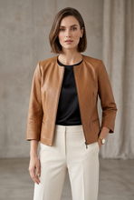 Įkelti nuotrauką į galerijos peržiūros programą, A woman modeling a cropped tan faux-leather jacket over a black top and cream trousers, standing against a neutral draped studio backdrop.
