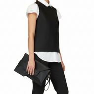 A model stands against a white studio background wearing a black sleeveless vest over a white short-sleeve collared shirt, black trousers, and taupe heeled ankle boots, while holding a black envelope-style clutch.