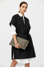 Load image into Gallery viewer, A woman models a black shift dress with a white collar and white undersleeves, holding a taupe envelope clutch bag. The background is a clean, neutral white studio backdrop.
