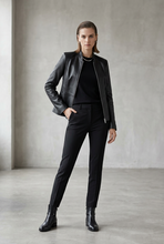 Load image into Gallery viewer, A woman modeling a black leather jacket, black top, black trousers, and black ankle boots in a minimalist studio setting with a textured gray wall and concrete floor.

