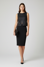 Load image into Gallery viewer, A woman models a black sleeveless midi dress with a structured peplum-style bodice, front zipper detail, and a matching black skirt. She is holding a small black clutch and wearing black pointed-toe heels against a light gray studio background.
