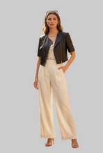 Įkelti nuotrauką į galerijos peržiūros programą, A woman modeling a black leather moto jacket with short sleeves, worn over a cream top and paired with cream wide-leg trousers, against a solid gray background.
