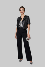 Įkelti nuotrauką į galerijos peržiūros programą, A woman models a black faux-leather cropped jacket with silver trim, paired with black wide-leg trousers and a patterned top, holding a silver clutch. She stands in a modern interior next to a gold wire Christmas tree.
