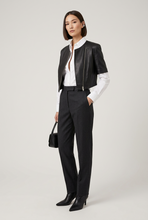 Įkelti nuotrauką į galerijos peržiūros programą, A model stands against a light gray studio background wearing a black leather moto jacket over a white collared shirt, paired with black trousers and black pointed-toe heels. She holds a small black handbag.

