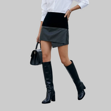 Load image into Gallery viewer, A woman wearing a black and gray mini skirt with a white long-sleeve top and black knee-high boots, holding a black handbag in an outdoor urban setting.
