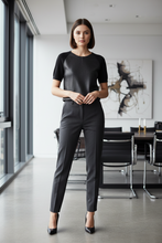 Load image into Gallery viewer, A woman stands in a modern office interior wearing a black short-sleeve top and dark charcoal gray trousers with black heels.
