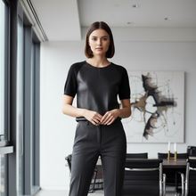 Load image into Gallery viewer, A woman stands in a modern office interior wearing a black short-sleeve top with a textured front panel and dark gray trousers.
