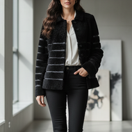 A woman walks down a modern, sunlit hallway wearing a black textured jacket with horizontal gray stripes, a white blouse, black skinny pants, and black ankle boots.