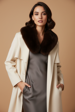 Įkelti nuotrauką į galerijos peržiūros programą, A woman modeling a long cream-colored coat with a large dark brown fur collar over a gray satin dress, set against a solid warm beige background.
