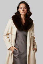 Įkelti nuotrauką į galerijos peržiūros programą, A woman modeling a long cream-colored coat with a large dark brown fur collar, worn over a dark gray satin dress, against a solid light gray studio background.
