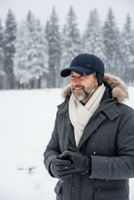 Įkelti nuotrauką į galerijos peržiūros programą, A man wearing a dark navy baseball cap, a thick cream knit scarf, a dark gray winter coat with a fur-trimmed hood, and black gloves, standing in a snowy outdoor setting with pine trees in the background.
