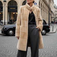 Įkelti nuotrauką į galerijos peržiūros programą, A woman stands on a Parisian street wearing a long beige patchwork faux-fur coat over a black top and dark gray trousers, with black sunglasses and a blurred black cab in the background.
