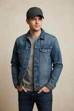 Įkelti nuotrauką į galerijos peržiūros programą, A man wearing a blue denim trucker jacket over a gray t-shirt, dark blue jeans, and a dark gray baseball cap, standing against a solid beige background.
