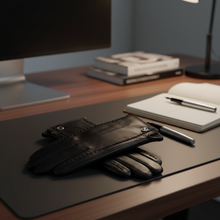 Įkelti nuotrauką į galerijos peržiūros programą, Black leather gloves with silver snap closures resting on a dark desk mat on a wooden desk, with a notebook, pen, books, and a computer monitor in the background.
