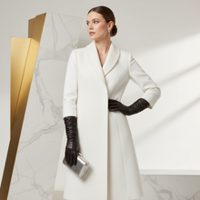 Įkelti nuotrauką į galerijos peržiūros programą, A woman models a long white coat with a shawl collar, paired with black gloves and holding a metallic silver clutch, set against a modern white marble and gold geometric background.
