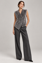 Load image into Gallery viewer, A model stands against a light gray studio background wearing a sleeveless wrap-style faux fur vest in heather gray over dark charcoal wide-leg trousers.
