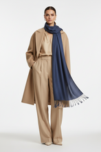 Įkelti nuotrauką į galerijos peržiūros programą, A model stands against a light gray studio background wearing a beige trench coat, matching wide-leg trousers, a beige blouse, and a dark navy blue scarf with fringe.
