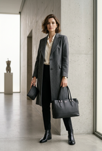 Load image into Gallery viewer, A woman stands in a modern concrete hallway wearing a long charcoal gray coat over a cream blouse and black skirt, holding a black leather tote bag and a small black quilted clutch.
