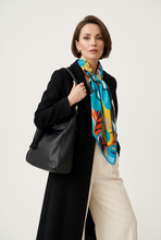 Įkelti nuotrauką į galerijos peržiūros programą, A woman models a vibrant multicolor silk scarf with a tropical leaf pattern, styled with a black coat and cream trousers while carrying a black leather shoulder bag against a plain light background.
