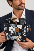 Load image into Gallery viewer, A man in a navy blazer holds a black floral zip pouch filled with grooming items, including a shaving brush, razor, and shaving cream. The pouch has a tropical leaf and flower print with a small tan brand patch reading Poma.
