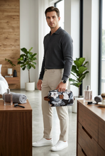 Įkelti nuotrauką į galerijos peržiūros programą, A man stands in a modern, sunlit bedroom holding a floral-patterned toiletry bag. He is wearing a dark gray polo shirt, beige trousers, and white sneakers.
