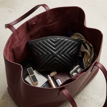 Įkelti nuotrauką į galerijos peržiūros programą, A burgundy leather tote bag shown open from above, filled with cosmetics and accessories including a black quilted pouch, a scarf, a lipstick, and earbuds case, on a light stone surface.
