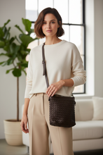 Įkelti nuotrauką į galerijos peržiūros programą, A woman wearing a cream knit sweater and beige trousers carries a dark brown woven crossbody bag with a single strap in a bright, modern living room.
