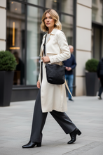 Įkelti nuotrauką į galerijos peržiūros programą, A woman walking on a city sidewalk wearing a long cream trench coat, dark wide-leg trousers, black ankle boots, and carrying a black quilted shoulder bag.
