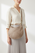 Įkelti nuotrauką į galerijos peržiūros programą, A woman wearing a cream button-up shirt and a light gray pleated skirt carries a woven beige crescent shoulder bag with a tan strap and brass hardware.
