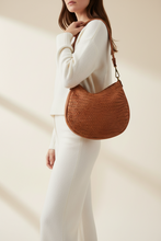 Įkelti nuotrauką į galerijos peržiūros programą, A woman wearing a cream knit sweater and matching pants carries a brown woven crescent shoulder bag with a long strap against a warm beige background.
