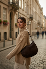 Įkelti nuotrauką į galerijos peržiūros programą, A woman wearing a beige knit turtleneck sweater and white trousers carries a dark brown woven shoulder bag on a cobblestone street with classic European buildings in the background.
