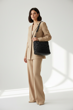 Įkelti nuotrauką į galerijos peržiūros programą, A woman modeling a beige two-piece suit and holding a black textured shoulder bag with a black strap, set against a bright white studio background with soft window-light shadows.
