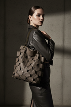 Įkelti nuotrauką į galerijos peržiūros programą, A model in a black leather outfit carries a large taupe woven leather tote bag with a gold chain strap against a textured concrete wall with dramatic side lighting.
