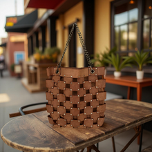 Load image into Gallery viewer, Brown woven handbag on a wooden table with a blurred street scene in the background
