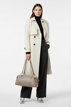 Įkelti nuotrauką į galerijos peržiūros programą, A model stands against a light gray studio background wearing a long cream double-breasted coat over a black turtleneck and black wide-leg trousers, while carrying a taupe leather handbag with silver-tone hardware.
