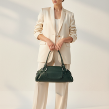 Įkelti nuotrauką į galerijos peržiūros programą, A model stands in a studio wearing a cream-colored blazer and matching trousers, holding a dark green leather handbag. The background is a warm off-white with soft diagonal shadows.
