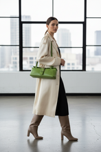 Įkelti nuotrauką į galerijos peržiūros programą, A woman walks indoors wearing a long cream coat, black dress, taupe knee-high boots, and carrying a bright green structured handbag with a top handle and flap detail.
