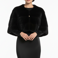 A model wearing a black long-sleeve faux-fur bolero jacket with a small gold button detail, paired with a matching black floor-length gown, against a plain white studio background.
