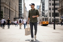 Įkelti nuotrauką į galerijos peržiūros programą, Man walking on a city street holding a tote bag, wearing a green shirt and black cap.
