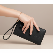 Įkelti nuotrauką į galerijos peržiūros programą, A black pebbled leather wristlet clutch with a matching wrist strap and gold-tone hardware, held by a person's hand against a neutral beige background.
