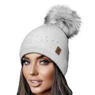 A white knitted cap with a grey artificial fur pom-pom and Swarovski crystals, featuring a small brown label with a brand logo displayed on a mannequin.