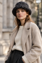 Load image into Gallery viewer, A woman wearing a chunky dark gray knit bucket hat, a cream ribbed knit sweater, and a beige wool-blend coat, photographed outdoors against a softly blurred stone building background.
