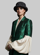 Load image into Gallery viewer, A model wearing a dark green brocade blazer with dramatic cream puff sleeves and a dark textured fur hat, set against a solid gray studio background.
