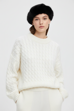 Įkelti nuotrauką į galerijos peržiūros programą, A model wearing a cream-colored cable-knit crewneck sweater, cream trousers, a black fur beret, and looking directly at the camera against a light gray studio background.
